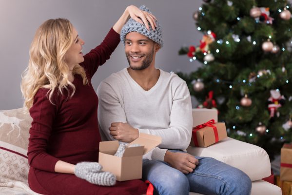 Happy woman putting on a tuque on her partner who looks a bit bewildered at his gift. Christmas Tree in background. Gifts for Men Blog vLife